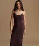 A woman modeling a dark plum-brown sleeveless slip dress with thin spaghetti straps and a subtle textured pattern, set against a solid warm beige studio background.