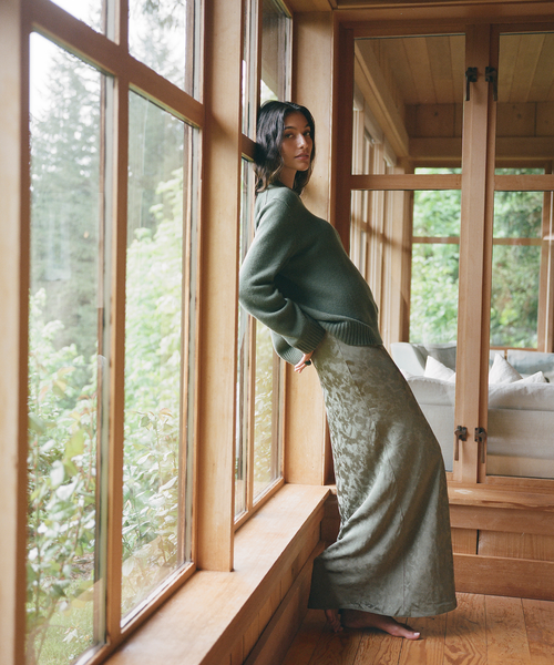 A woman stands barefoot by a large wooden window in a sunroom, wearing a muted sage green knit sweater and a matching textured sage green maxi skirt.