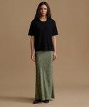A woman stands against a plain warm beige studio background wearing a loose black short-sleeve top and a long olive green skirt.