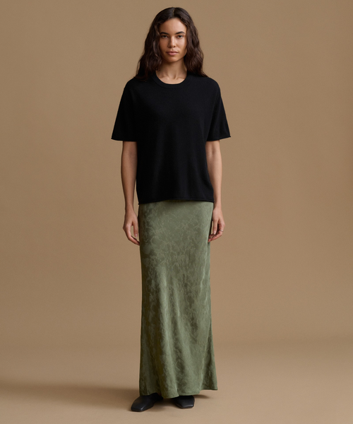 A woman stands against a plain warm beige studio background wearing a loose black short-sleeve top and a long olive green skirt.