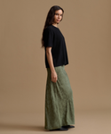 A woman stands in profile wearing a black short-sleeve top and a wide-leg sage green skirt against a plain beige studio background.