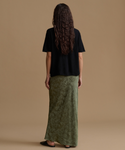 A woman is shown from behind wearing a black short-sleeve top and a long olive green patterned skirt against a solid tan studio background.