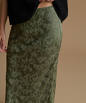 A close-up of a model wearing a high-waisted olive green skirt with a textured, distressed abstract pattern, paired with a black cropped top against a solid beige background.