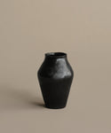 The JB1 Vase is made from glossy black ceramic, with a narrow neck and wide body, displayed on a neutral beige background. It has a simple matte finish and no visible decorations.