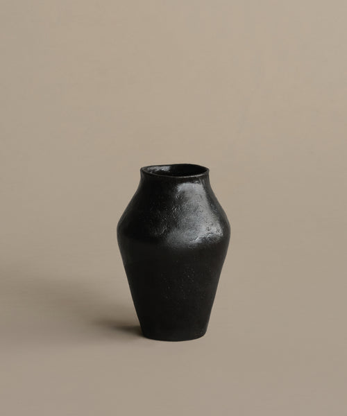 The JB1 Vase is made from glossy black ceramic, with a narrow neck and wide body, displayed on a neutral beige background. It has a simple matte finish and no visible decorations.