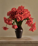 A glossy black JB1 Vase, filled with vibrant red double tulips, sits on a marble surface against a beige wall. Some tulip stems extend outward while others gently droop over the vase’s edge.