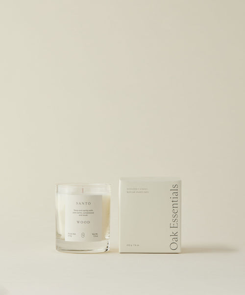 The Santo Wood Candle, featuring its signature glass design and woody scent, is displayed next to a white box labeled "Oak Essentials," all set against a simple light beige background.