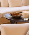 A lit Santo Wood Candle rests on a dark round table beside a bowl of dried botanicals, with cream couches in the background. Its musky scent brings warmth and an inviting feel to the cozy room.