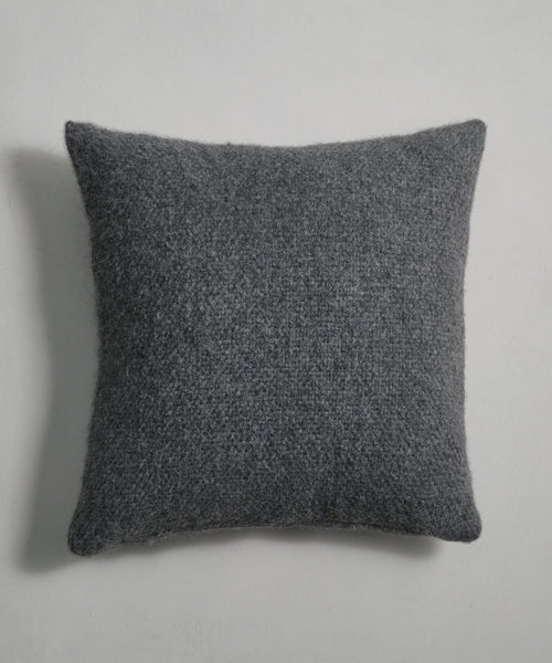 The Alpaca Basketweave Pillow, shown in dark gray with a square shape and slightly rounded corners, features a soft, fuzzy texture and is displayed against a plain light gray background.