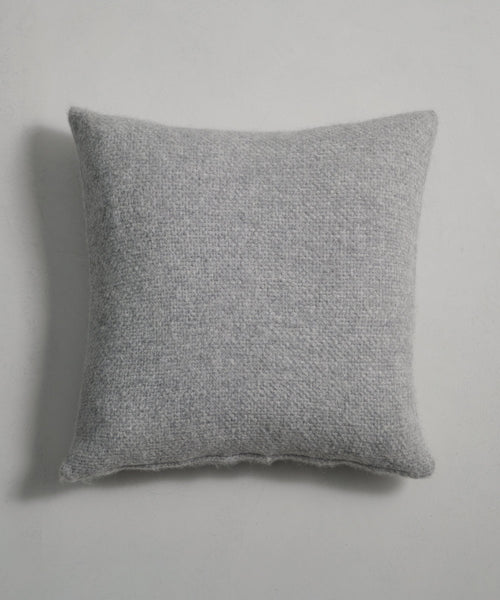 The Alpaca Basketweave Pillow in light gray rests on a plain, light surface, bringing cozy texture and style to the space.