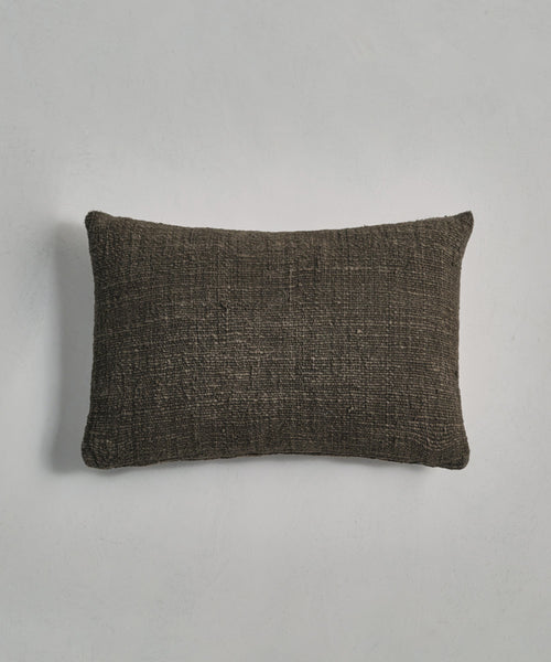 The Luna Lumbar Pillow, a rectangular dark brown handwoven pillow, is placed on a light gray surface.