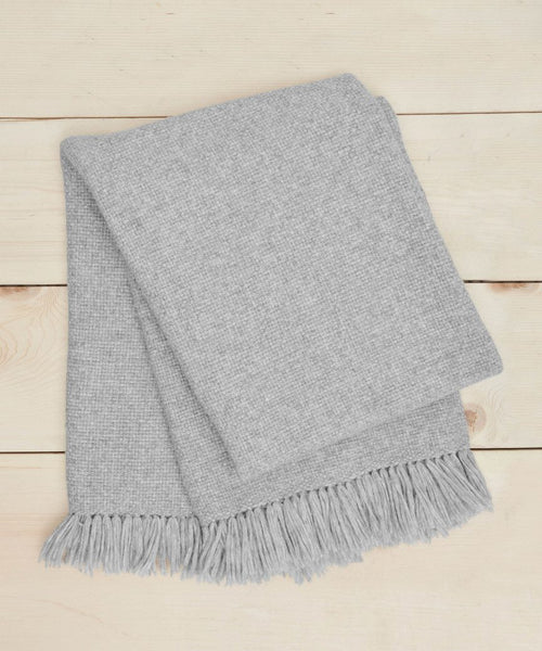 The Alpaca Basketweave Throw Rewards GWP, in light grey with fringed edges, is elegantly folded and displayed on a light wooden surface.