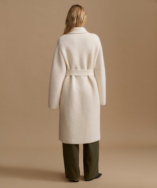 A woman viewed from the back wearing a long cream-colored belted coat over dark olive green trousers, set against a solid warm beige studio background.