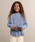 A young child with long curly hair smiles in a loose, light blue Kids Cashmere Fisherman sweater and cream pants, standing against a plain beige background.