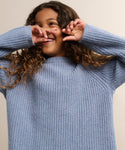 A child with curly brown hair smiles and playfully covers part of their face while wearing a light blue Kids Cashmere Fisherman sweater. The background is plain and neutral.
