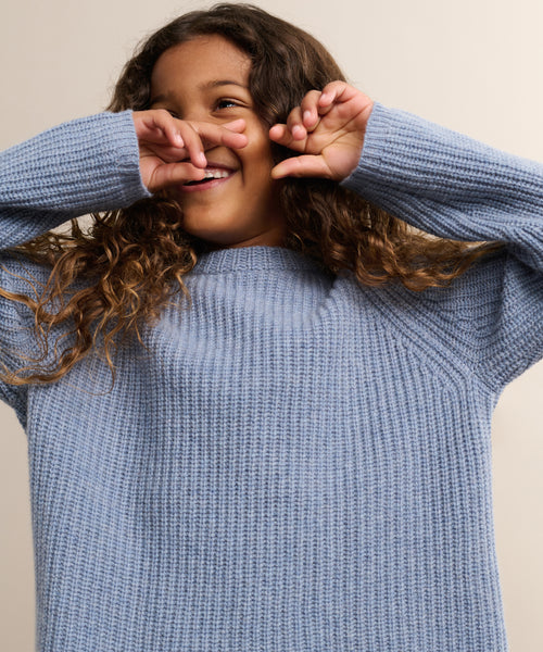 A child with curly brown hair smiles and playfully covers part of their face while wearing a light blue Kids Cashmere Fisherman sweater. The background is plain and neutral.