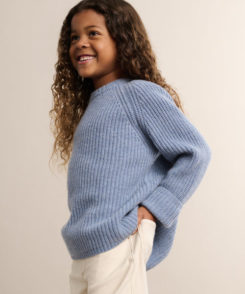 A smiling young child with long curly hair wears the Kids Cashmere Fisherman sweater in light blue and cream pants, posing with one hand in their pocket against a neutral backdrop.