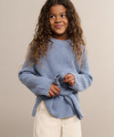 A young girl with long, curly hair smiles while looking to the side. She is wearing a light blue Kids Cashmere Fisherman sweater and light-colored pants, standing against a plain beige background.