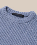 Close-up of the Kids Cashmere Fisherman sweater in light blue, featuring a ribbed crew neckline and a black label inside the collar, displayed against a beige background.