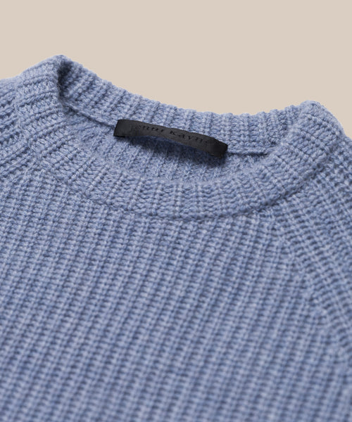 Close-up of the Kids Cashmere Fisherman sweater in light blue, featuring a ribbed crew neckline and a black label inside the collar, displayed against a beige background.
