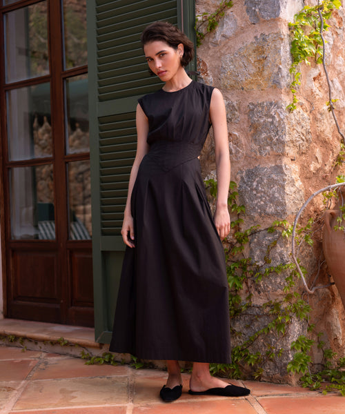 Wearing the sleeveless, long black Kit Dress with black flats, a woman stands calmly on a tiled patio beside a stone wall with green vines and wooden shutters, facing the camera.