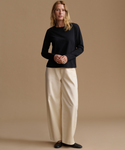 A woman stands against a plain warm beige studio background wearing a black long-sleeve top, cream wide-leg pants, and black flats.