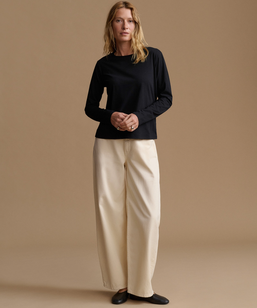 A woman stands against a plain warm beige studio background wearing a black long-sleeve top, cream wide-leg pants, and black flats.