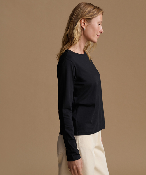 A woman in side profile wearing a black long-sleeve crewneck top and cream-colored pants, set against a solid warm beige studio background.