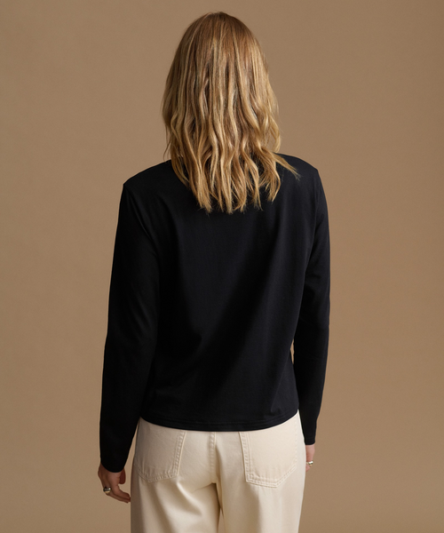 A woman viewed from behind wearing a black long-sleeve top and cream-colored pants, standing against a solid tan background.