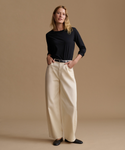 A woman stands against a warm beige studio background wearing a black long-sleeve top, cream wide-leg trousers, a thin dark belt, and black flats.