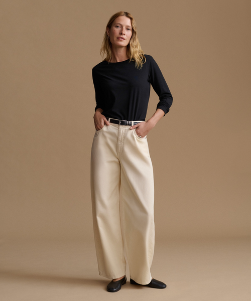 A woman stands against a warm beige studio background wearing a black long-sleeve top, cream wide-leg trousers, a thin dark belt, and black flats.