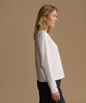 A woman in side profile wearing a long-sleeve white blouse and dark pants, standing against a solid beige studio background.
