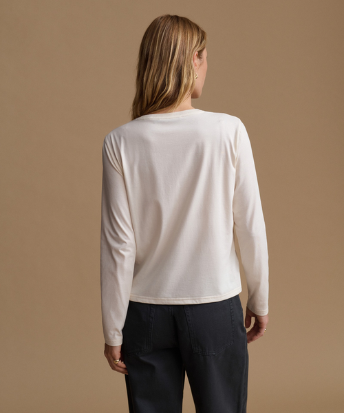 A woman shown from the back wearing a light cream long-sleeve top and dark charcoal pants, standing against a solid warm beige studio background.