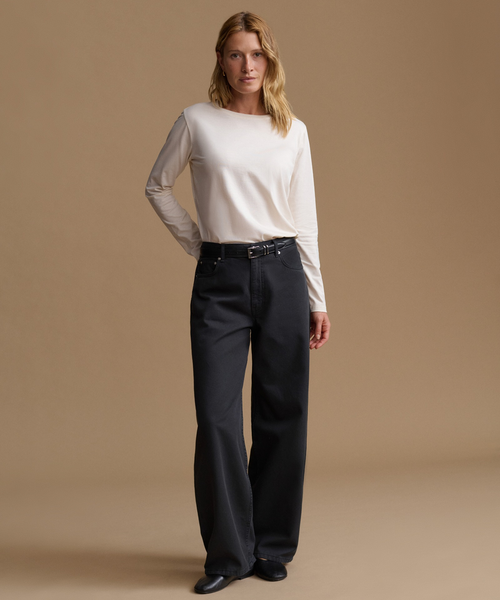 A woman stands against a plain beige studio background wearing a cream long-sleeve top, dark charcoal wide-leg pants, and black shoes.
