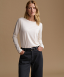A woman modeling a long-sleeve cream knit top tucked into dark charcoal pants with a black belt, set against a solid warm beige studio background.
