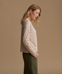 A woman in side profile wearing a light beige long-sleeve crewneck top and olive green pants, standing against a solid warm beige studio background.