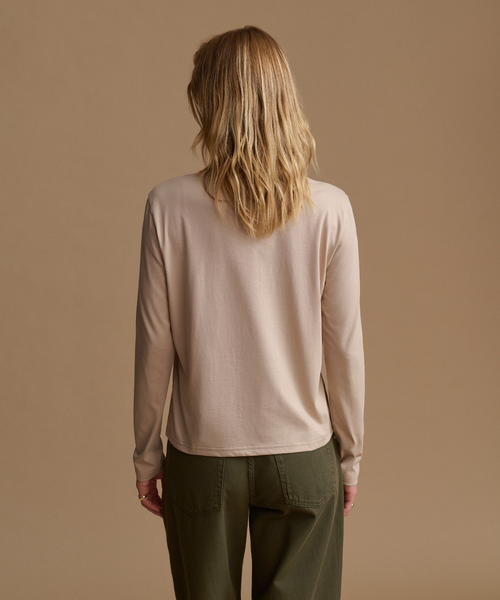 A woman viewed from behind wearing a light beige long-sleeve knit top and olive green pants, standing against a solid warm beige studio background.