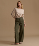 A woman stands against a warm beige studio background wearing a light beige long-sleeve top, olive green wide-leg pants, and taupe flats.