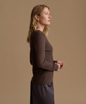 A woman in side profile wearing a fitted, long-sleeve ribbed knit top in dark brown, paired with a dark skirt, set against a solid warm beige studio background.