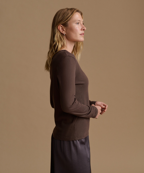 A woman in side profile wearing a fitted, long-sleeve ribbed knit top in dark brown, paired with a dark skirt, set against a solid warm beige studio background.