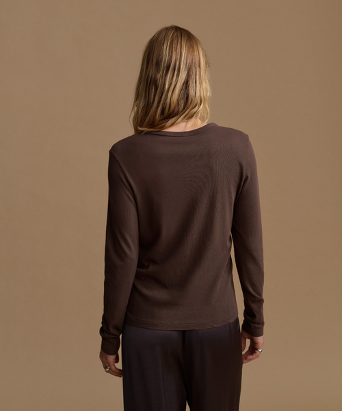 A woman viewed from behind wearing a dark brown ribbed long-sleeve top and dark pants against a solid tan studio background.