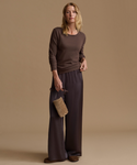 A woman stands against a warm beige studio background wearing a dark brown long-sleeve knit top and matching wide-leg trousers, holding a small textured tan clutch.