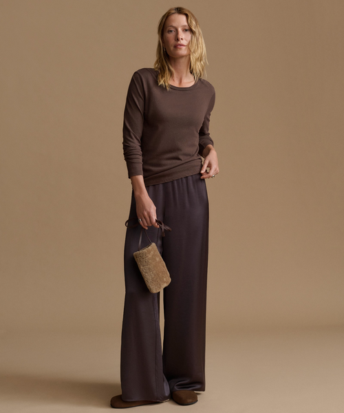 A woman stands against a warm beige studio background wearing a dark brown long-sleeve knit top and matching wide-leg trousers, holding a small textured tan clutch.
