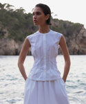 A woman in a sleeveless white Lara Top and matching skirt stands with her hands in her pockets by the water, rocky cliffs and trees in the background.
