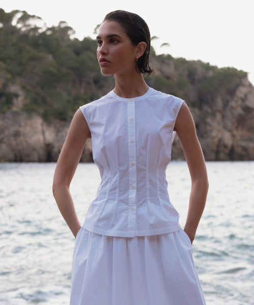 A woman in a sleeveless white Lara Top and matching skirt stands with her hands in her pockets by the water, rocky cliffs and trees in the background.