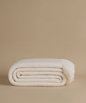 A folded cream-colored knit throw blanket with fringed ends, displayed on a solid beige studio background.