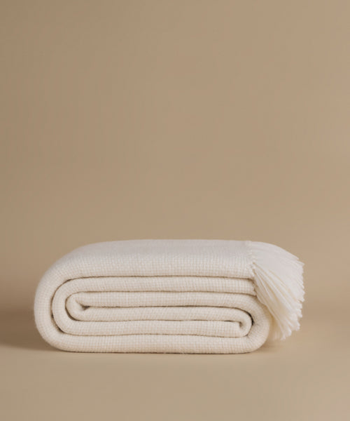 A folded cream-colored knit throw blanket with fringed ends, displayed on a solid beige studio background.