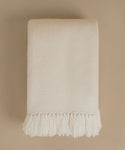 A folded cream-colored woven throw blanket with fringed edges, displayed against a solid beige background.