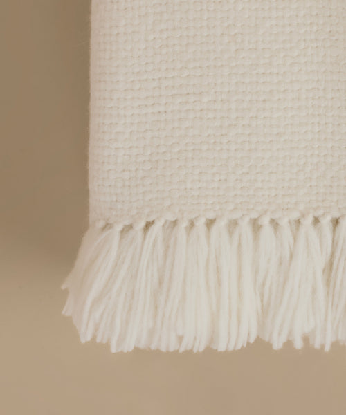 A folded cream-colored woven throw blanket with fringed ends, displayed against a solid beige background.