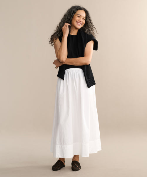 A woman with long curly hair smiles against a beige background. She wears a black sleeveless top, the wardrobe staple white Lauren Skirt—an ankle-length cotton style—and dark woven shoes.